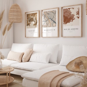 May include: Three framed city map prints in neutral tones hang above a white sofa. The prints feature the names Santos, Mannheim, and Rio de Janeiro. The room is decorated with a woven pendant light, dried plants, and a straw hat.