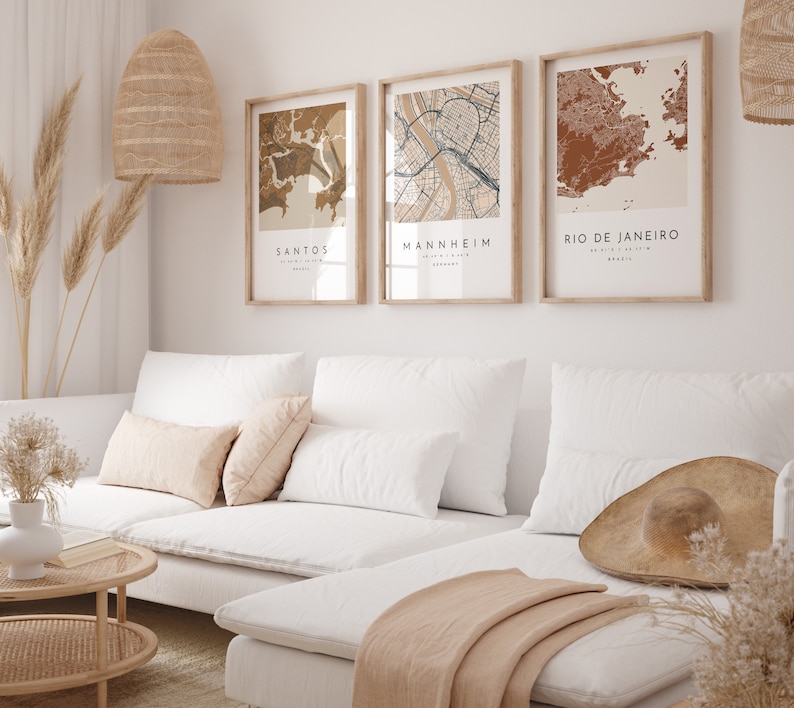 May include: Three framed city map prints in neutral tones hang above a white sofa. The prints feature the names Santos, Mannheim, and Rio de Janeiro. The room is decorated with a woven pendant light, dried plants, and a straw hat.