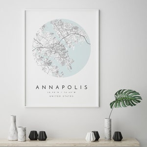 May include: A framed print of a map of Annapolis, Maryland, with a light blue background and black lines. The text "ANNAPOLIS" is printed in black below the map, along with the coordinates "38.98&deg;N / 76.49&deg;W" and "UNITED STATES".