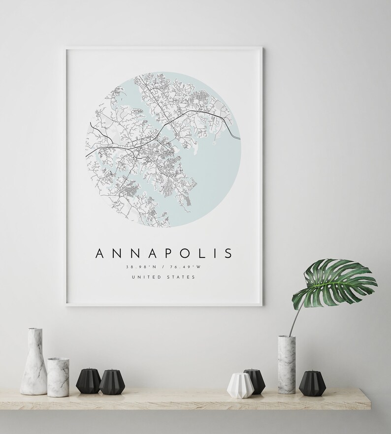May include: A framed print of a map of Annapolis, Maryland, with a light blue background and black lines. The text "ANNAPOLIS" is printed in black below the map, along with the coordinates "38.98&deg;N / 76.49&deg;W" and "UNITED STATES".