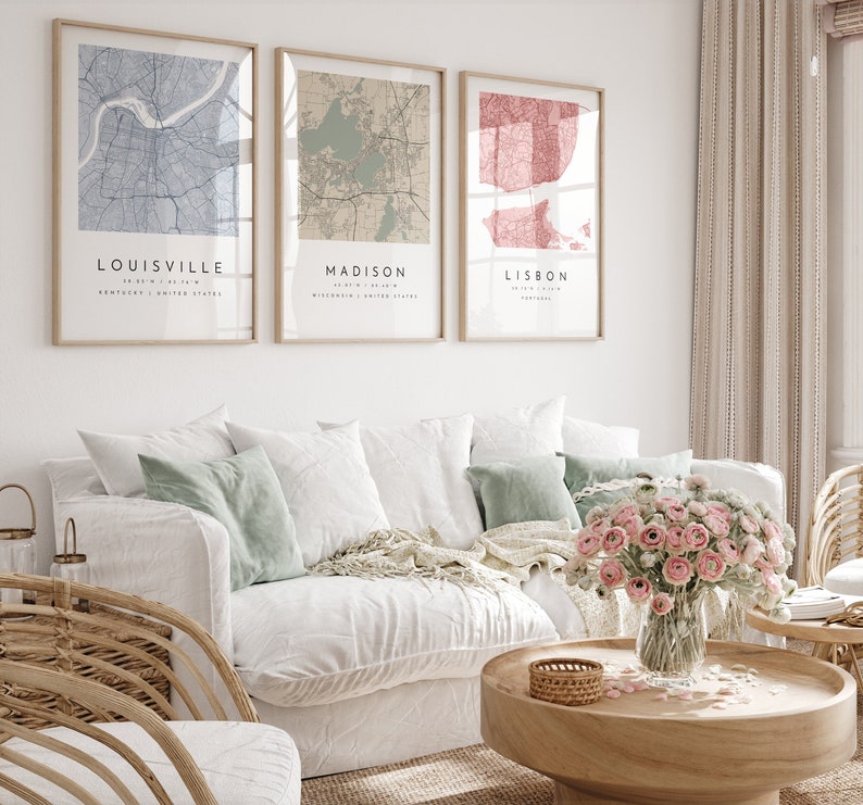 May include: Three framed city map prints. The maps are of Louisville, Madison, and Lisbon. The Louisville map is blue, the Madison map is tan, and the Lisbon map is red. The prints are displayed above a white couch with decorative pillows and a wooden coffee table.