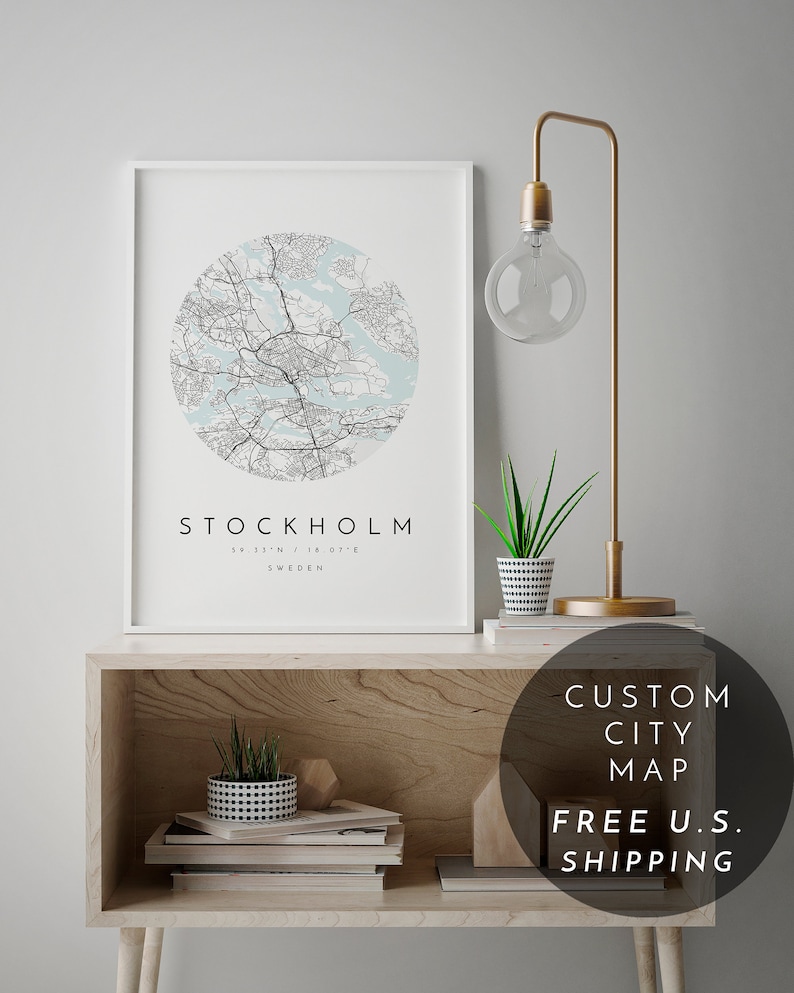 May include: A white framed print of a black and white city map of Stockholm, Sweden. The map is in a circle with a light blue background. The print is on a wooden shelf with a gold lamp and two potted plants.