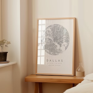 Puede incluir: Un mapa enmarcado de Dallas, Texas, en un estilo minimalista. El mapa es un diseño circular con una red de calles y carreteras grises. El nombre de la ciudad y las coordenadas están impresos debajo del mapa en una fuente moderna.