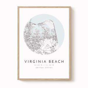 May include: A framed print featuring a detailed map of Virginia Beach, Virginia, within a light blue circular background. The city name and coordinates are printed below the map in a minimalist style. The frame is made of light-colored wood.