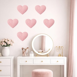 Könnte beinhalten: Ein hellrosa Schminktisch-Set mit rundem Spiegel, weißer Kommode und rosa gepolstertem Hocker. Sechs rosa gestrickte Herzdekorationen befinden sich an der Wand. Eine Vase mit rosa Blumen steht auf der Kommode.