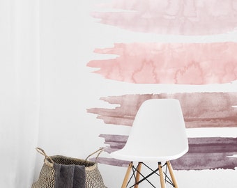 May include: Four horizontal watercolor brushstrokes in shades of pink and brown on a white wall. The brushstrokes are arranged in a gradient from light pink to dark brown. A white chair with wooden legs is in the foreground.