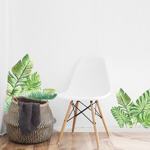 May include: A white chair with wooden legs and a woven basket with a dark gray blanket. Watercolor-style green banana leaf wall decals are on the white wall, creating a tropical theme.