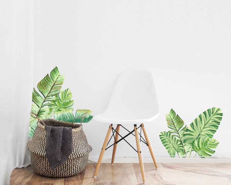 May include: A white chair with wooden legs and a woven basket with a dark gray blanket. Watercolor-style green banana leaf wall decals are on the white wall, creating a tropical theme.
