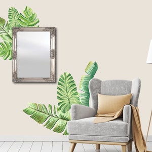 May include: A silver framed mirror hangs on a beige wall, adorned with watercolor-style green tropical leaves. A gray armchair with a yellow pillow and a brown throw blanket sits below the leaves. A wooden floor lamp with a white shade is to the right.
