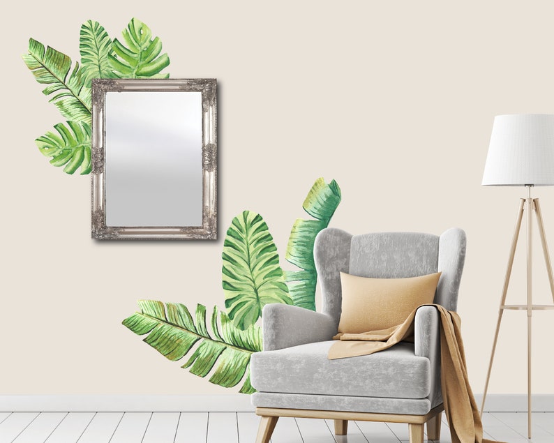 May include: A silver framed mirror hangs on a beige wall, adorned with watercolor-style green tropical leaves. A gray armchair with a yellow pillow and a brown throw blanket sits below the leaves. A wooden floor lamp with a white shade is to the right.