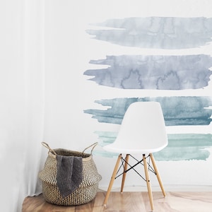 May include: Four horizontal watercolor brushstrokes in shades of blue and green on a white wall. The brushstrokes are arranged in a graduated pattern, with the darkest blue at the top and the lightest green at the bottom. A white chair with wooden legs sits in front of the wall.