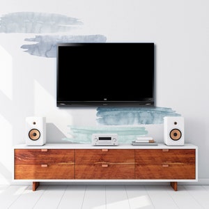 May include: A wooden entertainment center with four drawers and a white top. A flat screen TV is mounted on the wall above the entertainment center. Two white speakers with gold accents are on either side of the entertainment center. The wall behind the entertainment center has a light blue and green watercolor design.