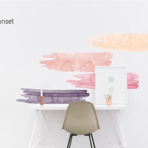 May include: A white wall with four watercolor brushstrokes in shades of pink, orange, and purple. The brushstrokes are arranged in a staggered pattern. A white desk with a white chair, a picture frame, and a small plant are in the foreground. The text "Sunset" is at the top of the image.