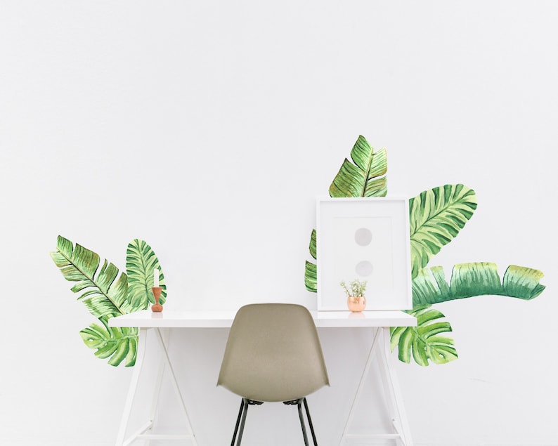May include: A white desk with a minimalist design, a gray chair, and a white picture frame. The wall is decorated with large, green watercolor banana leaf decals. A small copper vase and a wooden candle holder are on the desk.