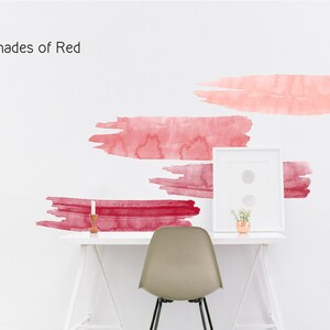 May include: Shades of Red wall decals featuring four abstract watercolor brushstrokes in various shades of red, pink, and burgundy. The decals are applied to a white wall above a white desk with a chair and a small potted plant.