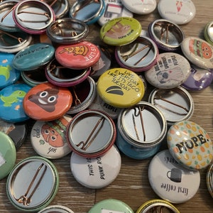 May include: A collection of colorful button pins with various designs and sayings. The pins are arranged on a wooden surface. Some of the designs include a poop emoji, a taco, a dinosaur, and a sloth. The text on the pins includes phrases like "Live Every Day Like It's Taco Tuesday", "This is Punk Rock", and "First Coffee".