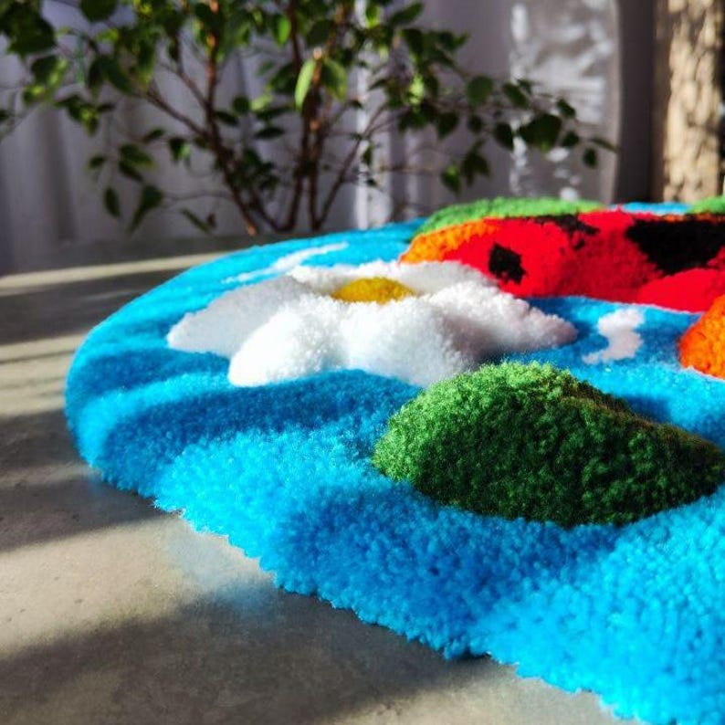 May include: A tufted rug featuring a vibrant blue background with a white daisy flower, a green hill, and a red ladybug with black spots. The textile surface has a thick, plush pile texture with distinct raised shapes and colourful yarn details.