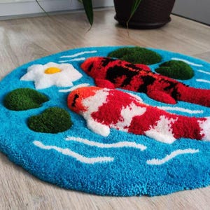 May include: A round tufted rug featuring a bright blue background with white ripples, two koi fish in red, white, and black, several dark green lily pads, and a single white water lily flower with a yellow centre, all rendered in a textured pile.