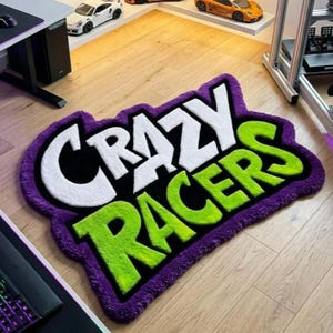 May include: A rectangular rug with the words "CRAZY RACERS" in white and green, outlined in black and purple. The rug is on a wooden floor, with toy cars in the background.