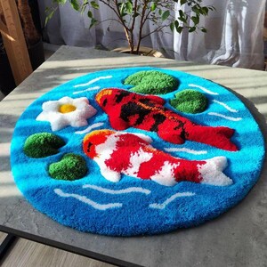 May include: A circular tufted rug featuring a vibrant blue water background with white ripple accents. The design includes two koi fish, one primarily red and black and the other red and white, alongside a white water lily and green lily pads.