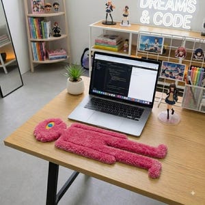 May include: A pink, fuzzy, tufted rug shaped like a stylized human figure with a single yellow and blue eye detail on the head. The rug is positioned on a wooden desk in front of an open laptop displaying code, alongside a small potted plant.