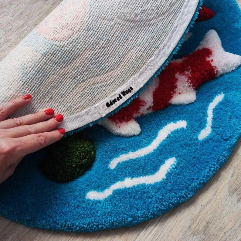 May include: A round tufted rug featuring a blue background with white wavy lines and a red and white koi fish design. The underside shows a white mesh backing with a fabric border label that reads Adored Rugs in black text.