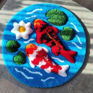 May include: A circular tufted rug featuring a vibrant blue water background with white ripple accents. The design includes two koi fish, one primarily red and black and the other white and orange, alongside a white water lily and green lily pads.