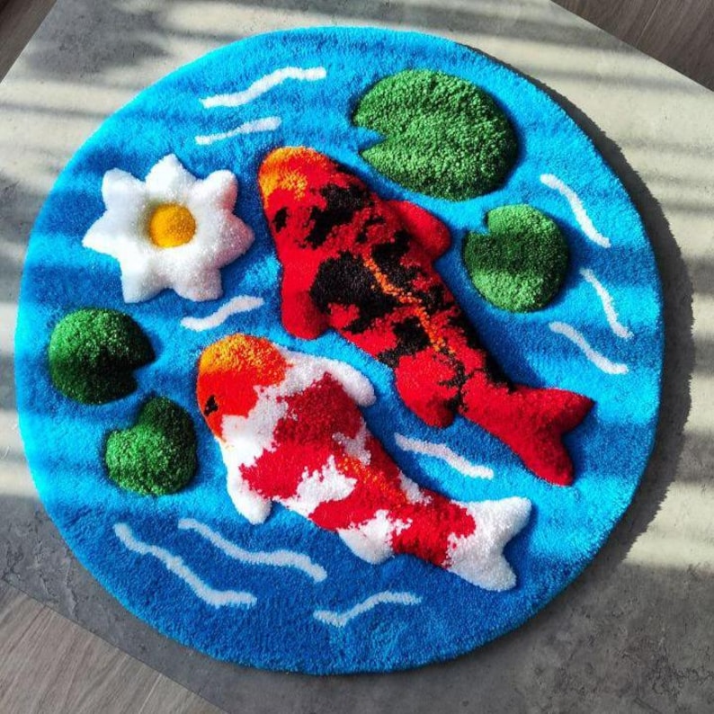 May include: A circular tufted rug featuring a vibrant blue water background with white ripple accents. The design includes two koi fish, one primarily red and black and the other white and orange, alongside a white water lily and green lily pads.