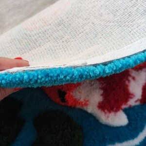 May include: A tufted rug with a thick, bright blue border and a white mesh backing. The visible surface features a pattern with red, white, and black tufted fibres, creating a textured, plush appearance with a distinct edge showing the construction.