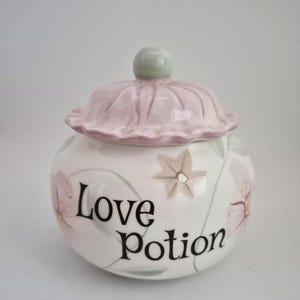 May include: A round ceramic jar with a lid, featuring a pale pink ruffled top and a light green knob. The white body is decorated with painted pink flowers and green leaves, with the black text Love Potion printed on the front surface.