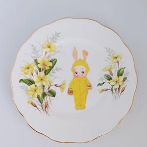 May include: A white ceramic plate with a scalloped edge and gold trim. The plate features a cartoon illustration of a yellow bunny character holding a basket, surrounded by yellow flowers and green leaves.