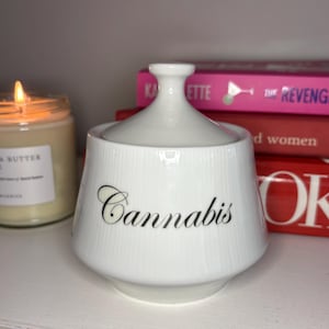 May include: A white ceramic jar with a matching lid, featuring a subtle vertical ribbed texture. The word Cannabis is printed in a black cursive script on the front of the jar. The container has a wide base that tapers towards the top.