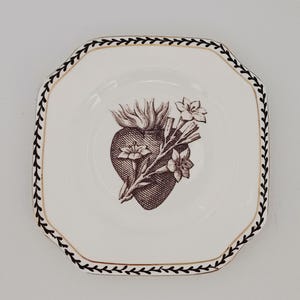 May include: A white, octagonal ceramic plate featuring a brown heart and floral design in the centre. The plate has a black and gold leaf border. A decorative item.