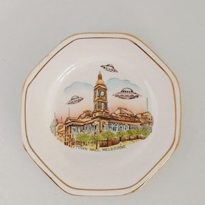 May include: A small, octagonal decorative plate with a gold rim. The plate features a painted scene of a building with a clock tower, three UFOs, and the text "TOWN HALL MELBOURNE". The plate has a cream-coloured background.