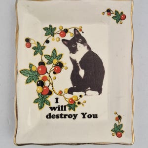 May include: A rectangular ceramic trinket tray with a gold-trimmed scalloped edge. The centre features a black and white cat illustration, a berry vine graphic, and the black text "I will destroy You" printed on a cream-coloured background.