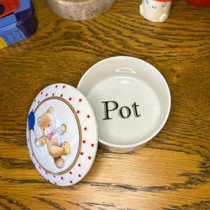 May include: A white ceramic round trinket box with a removable lid. The lid features a brown teddy bear holding a blue balloon, surrounded by a red polka dot border. The interior of the base has the word Pot printed in black serif font.