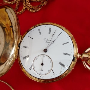 May include: A gold-tone pocket watch with an open case, featuring a white dial, black Roman numerals, and a sub-dial for seconds. The watch includes a matching gold-tone chain and displays the text L. Jacot Locle on the face of the dial.