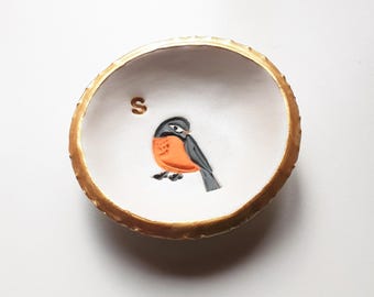 Könnte beinhalten: Eine kleine, weiße Keramikschale mit goldenem Rand. Die Schale zeigt eine gemalte Illustration eines Vogels mit orangefarbener Brust, grauen Flügeln und einem schwarzen Kopf. Der Buchstabe "S" ist über dem Vogel gemalt.