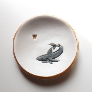 May include: White ceramic ring dish with a gold rim and a grey shark illustration. The letter "W" is written in gold on the dish.