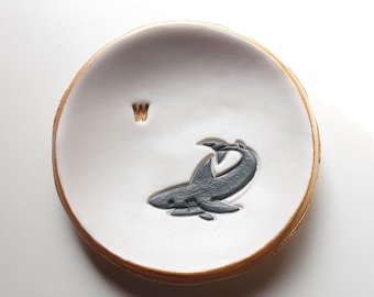 May include: White ceramic ring dish with a gold rim and a gray shark illustration. The letter "W" is written in gold on the dish.