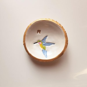 Puede incluir: Un pequeño plato de cerámica blanca con un borde dorado, que presenta un colibrí pintado a mano en vuelo. El plato tiene la letra "R" pintada en el interior.