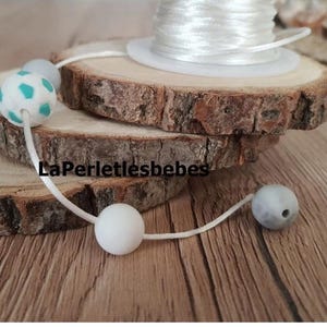 Peut inclure: Un cordon blanc avec une perle en silicone turquoise et blanche, une perle en silicone blanche et deux perles en silicone grises. Les perles sont enfilées sur le cordon blanc. Le cordon est sur une surface en bois.