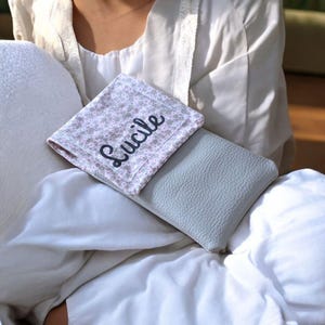 Puede incluir: Una funda de cuero gris claro con un cuadrado de tela estampada floral encima. El cuadrado de tela tiene el nombre "Lucile" bordado en escritura negra. La funda y la tela descansan sobre una superficie blanca.