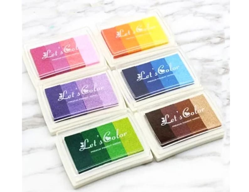 May include: Six colorful ink pads with the text "Let's Color" on each pad. The ink pads are arranged in two rows of three. The ink pads are in the colors of orange, pink, purple, blue, brown, and green.
