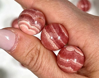 Könnte beinhalten: Drei rosa und weiße, gestreifte Kugeln aus rosa Calcit, auch bekannt als mexikanischer rosa Calcit, werden in einer Hand gehalten. Die Kugeln sind glatt und poliert.