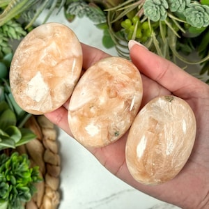 Op de afbeelding: Drie gladde, ovale, oranje en witte seleniet palmstenen. De stenen hebben een natuurlijke, gepolijste afwerking.