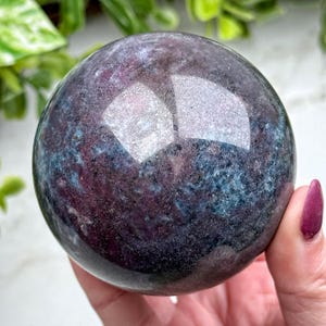 May include: A polished, spherical gemstone with a mix of deep purple and teal colors. The orb has a reflective surface, and is held in a person's hand. The gemstone is a decorative item.