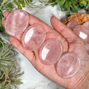 May include: Six smooth, oval-shaped rose quartz stones, each with a pale pink color and a translucent finish. The stones are arranged in a hand, with the largest stone at the top and the smallest stone at the bottom.