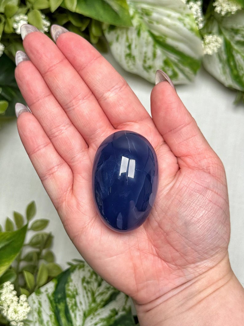 May include: A smooth, oval-shaped, deep blue gemstone. The polished stone is held in a hand, with green and white foliage in the background. The stone's surface reflects light, highlighting its glossy finish.