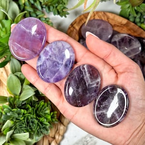 May include: A hand holds several polished amethyst stones. The stones are oval-shaped and display a range of purple hues, from light lavender to deep violet, with natural veining patterns. The stones are smooth and reflective, suggesting a high polish.